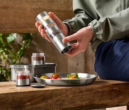 Une personne assaisonne un plat avec un moulin à épices en acier inoxydable. Sur la table, il y a d'autres récipients à épices et un plat de nourriture.