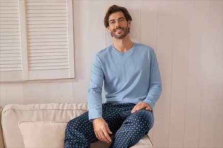 Un homme souriant avec une barbe est assis sur un canapé clair, portant un t-shirt bleu clair à manches longues et un pantalon de pyjama bleu foncé à motifs. Des volets de fenêtre blancs sont visibles en arrière-plan.