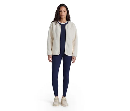 Femme debout, portant une veste polaire crème sur un haut et un legging bleu marine, avec des baskets claires.