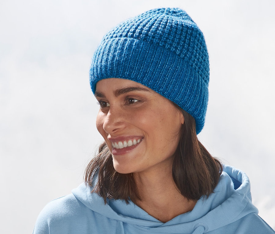 Une femme les bras croisés devant une montagne porte un bonnet bleu en maille.