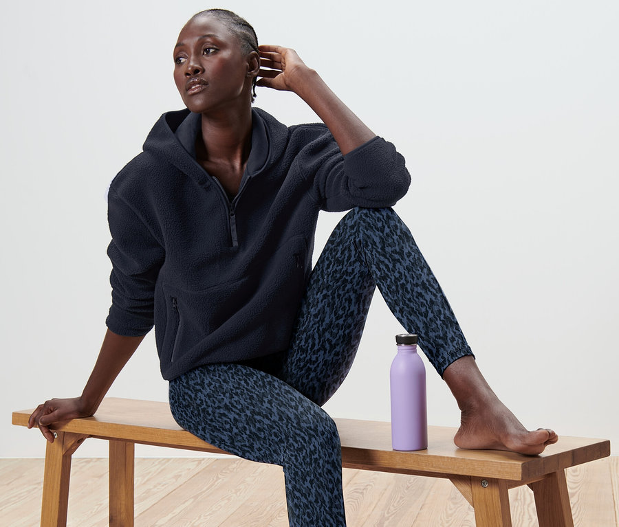 Femme assise sur un banc portant un pull à capuche en peluche teddy et des leggings à motifs, avec une bouteille violette à côté d'elle.