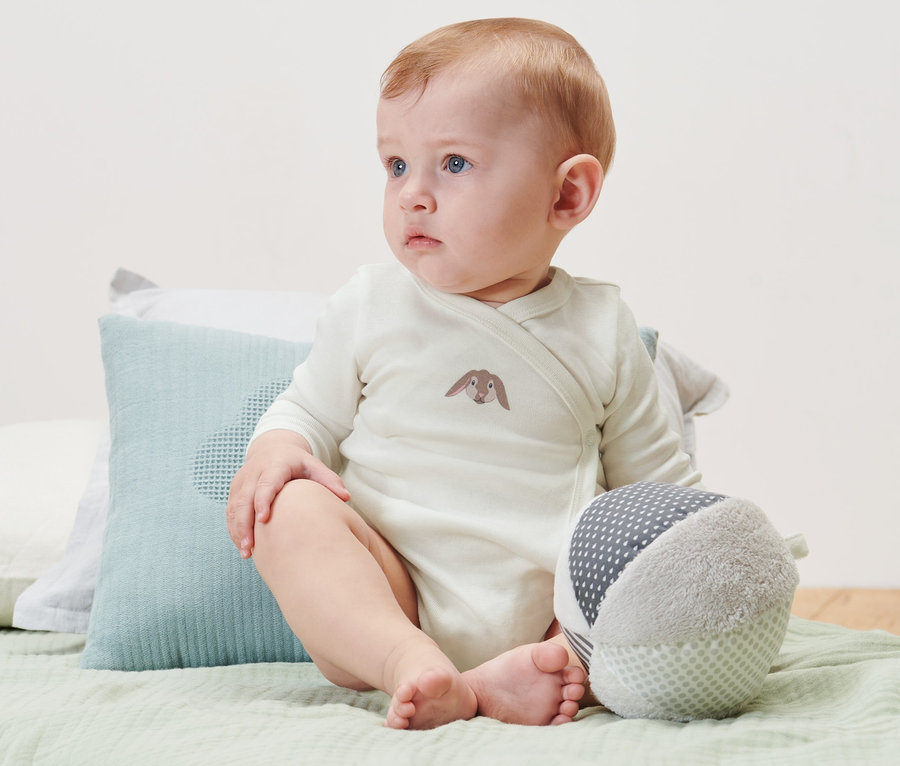 Un bébé est assis sur un lit et porte 3 bodys à manches longues pour bébé.