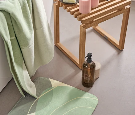 Tapis de bain confortable vert, linge de bain XL aux formes organiques et support avec distributeur de savon près de la baignoire. Sur un tabouret en bois se trouvent 2 bougies à LED en cire véritable et un livre ouvert.