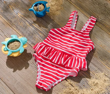 Un maillot de bain pour enfant rayé rouge et blanc est posé sur une surface en bois à côté de deux jouets de sable.