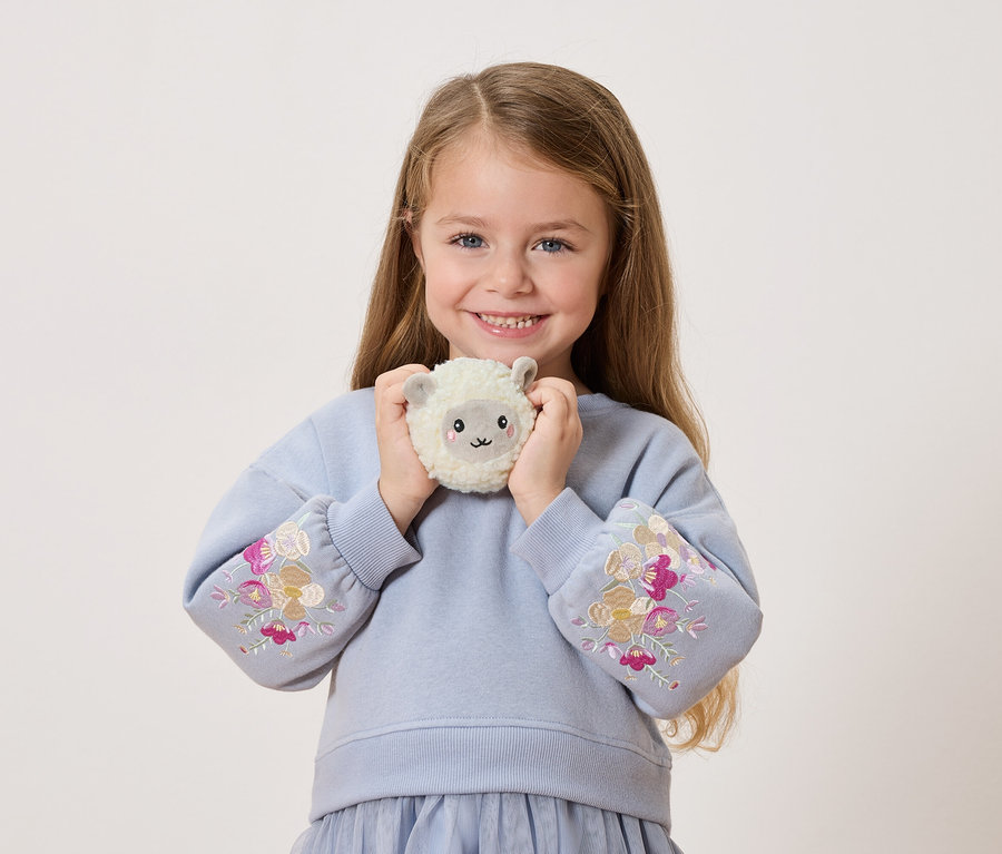 Une jeune fille souriante tient un mouton en peluche devant elle. Elle porte un pull bleu avec des broderies florales sur les manches.