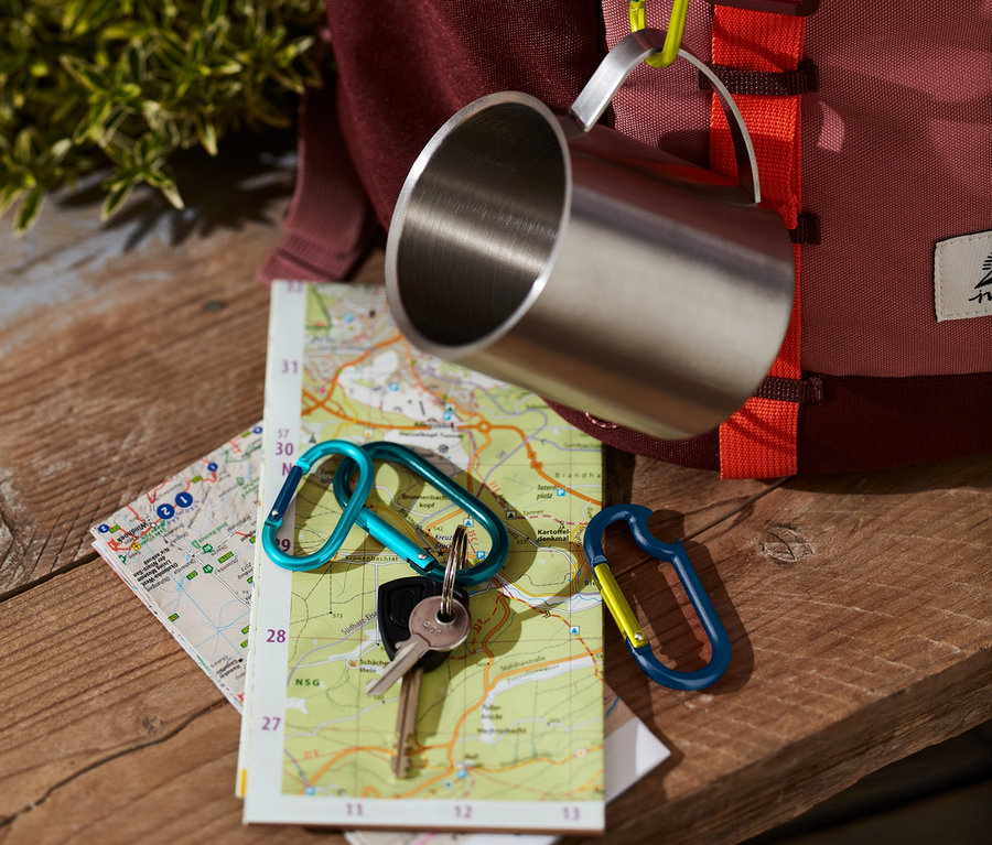 Une tasse en acier inoxydable est accrochée à un sac à dos à l'aide d'un des 4 mousquetons. Des clés avec un autre mousqueton reposent sur une carte.