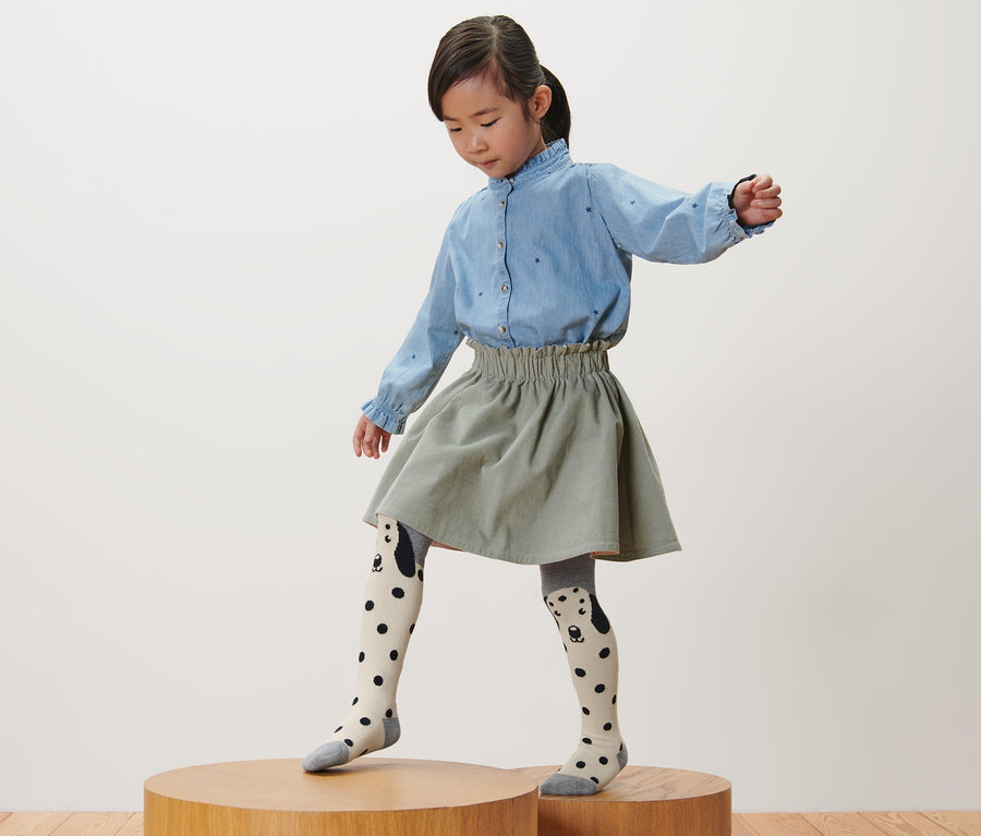 Une fille se tient sur un socle en bois et porte 3 collants pour enfant, rose, gris, rose baies.