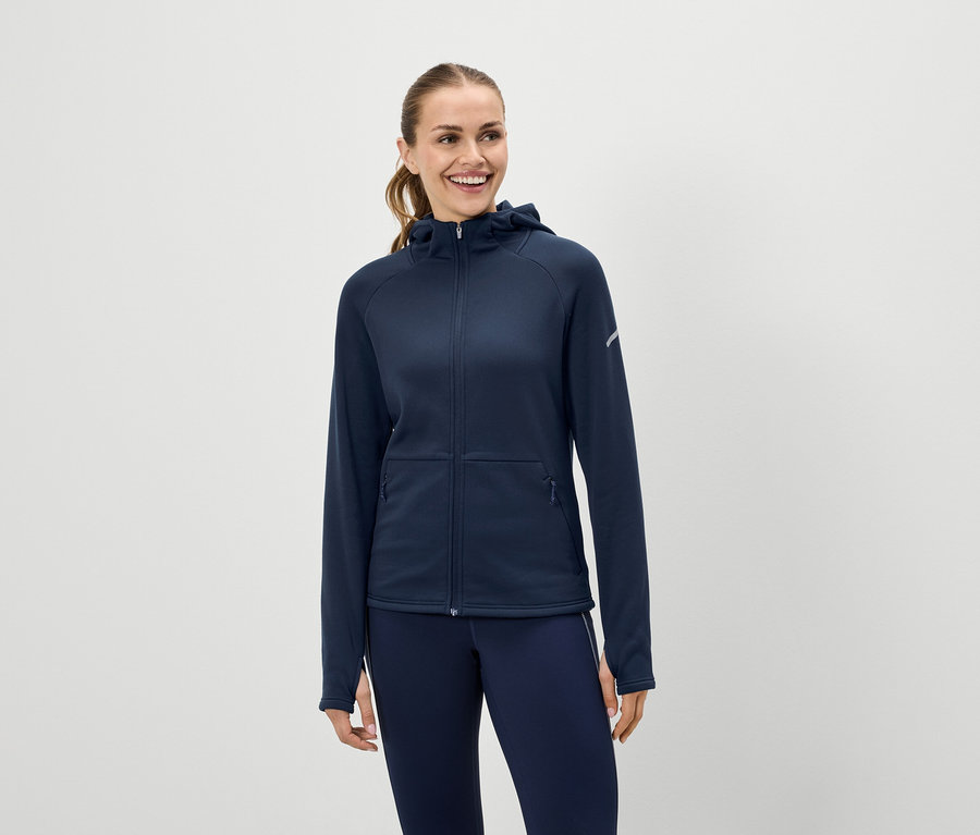 Une femme souriante porte un sweat à capuche bleu marine zippé et un legging bleu marine.