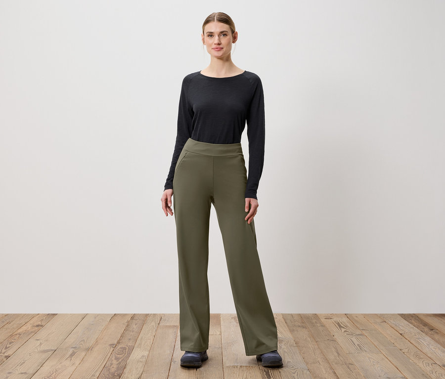 Une femme pose sur un plancher en bois, portant un pantalon vert olive et un haut noir à manches longues.