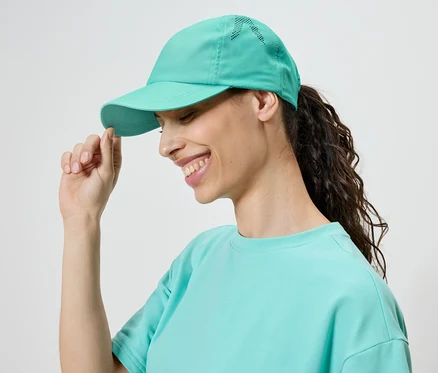 Une femme souriante portant une casquette et un t-shirt turquoise touche la visière de sa casquette.