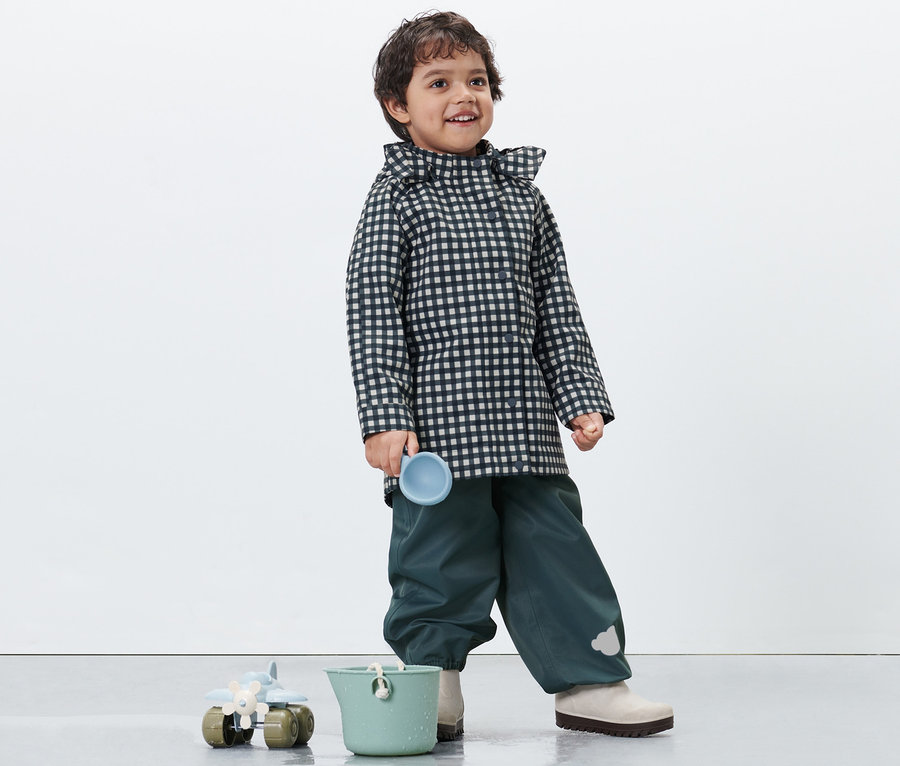 Un garçon portant un imperméable pour tout petit à carreaux tient une tasse bleue. Un seau vert et un jouet sont en bas.
