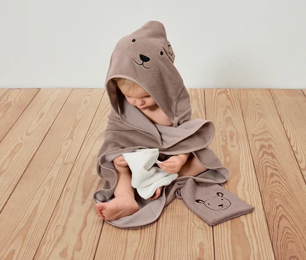 Un enfant est assis sur un plancher en bois, enveloppé dans une serviette à capuche marron avec un motif d'ours et tenant un chiffon blanc.