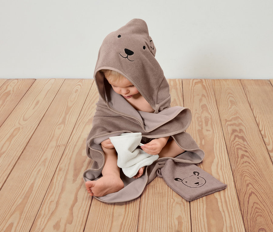 Un enfant est assis sur un plancher en bois, enveloppé dans une serviette à capuche marron avec un motif d'ours et tenant un chiffon blanc.