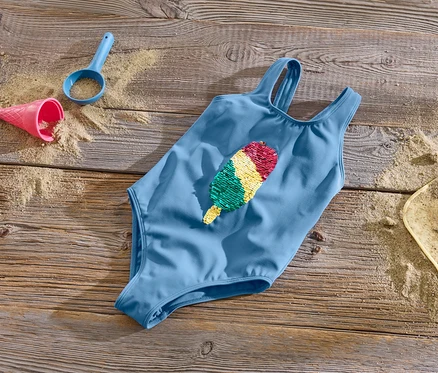 Un maillot de bain bleu pour enfant avec des paillettes réversibles est posé sur une surface en bois avec du sable et des jouets.