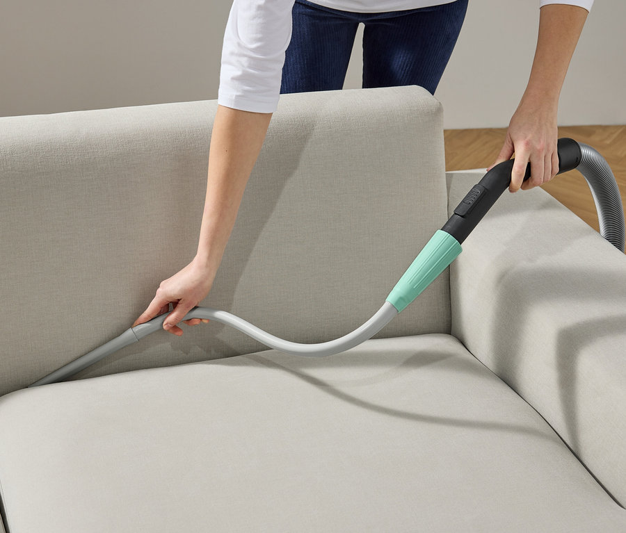 Une femme nettoie un canapé avec un embout d'aspirateur.