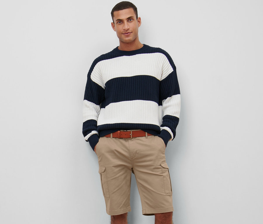 Un homme pose en short cargo beige et pull à rayures marine et blanches.
