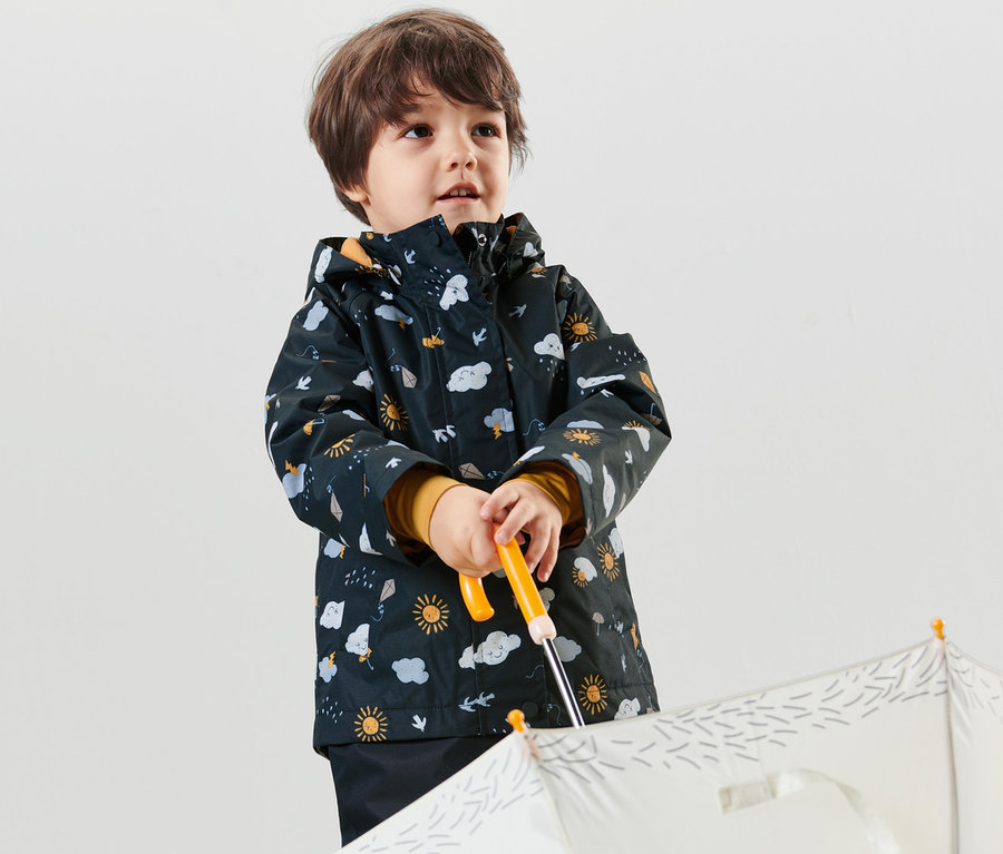 Un jeune garçon tient un parapluie blanc et porte une veste de pluie thermique bleu foncé imprimée intégrale.