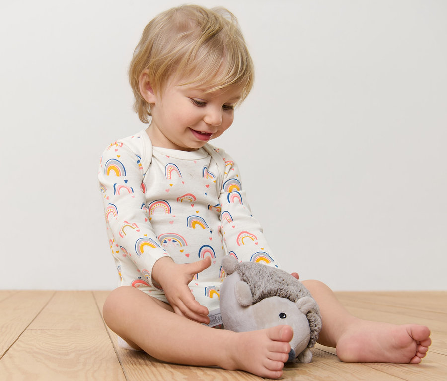 Un enfant est assis par terre et joue avec un hérisson en peluche. Il porte un body à manches longues avec un imprimé arc-en-ciel.