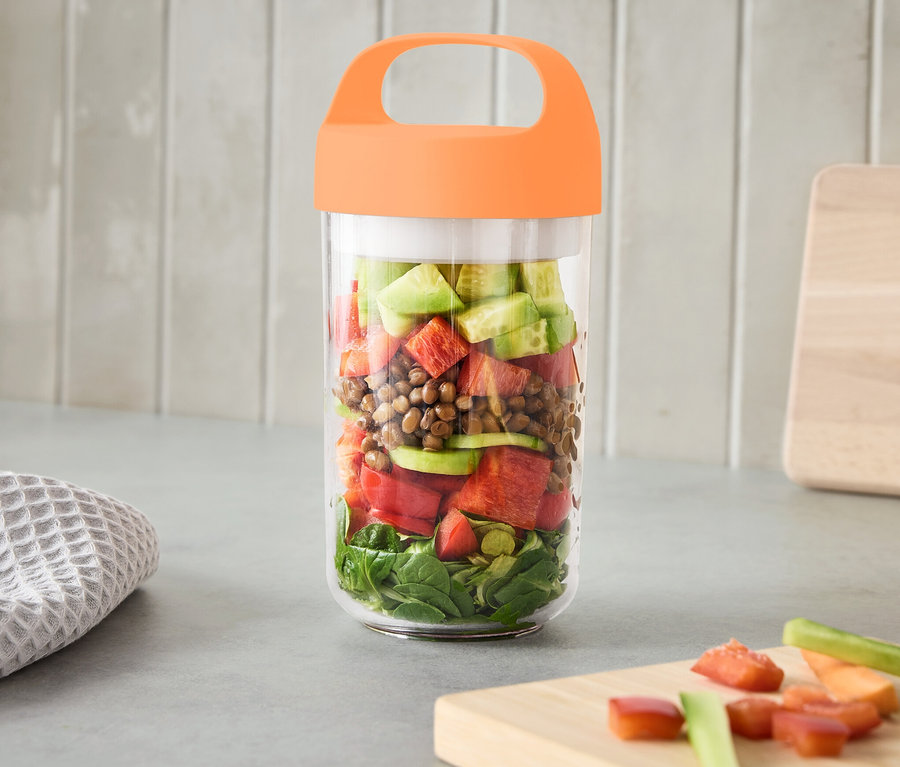 Un grand récipient pour produits à emporter avec poignée, rempli de légumes, est posé sur un comptoir de cuisine.