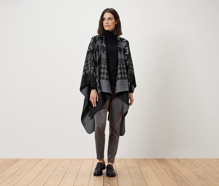 Une femme debout portant un col roulé noir, un pantalon gris et une cape pied-de-poule grise et noire.