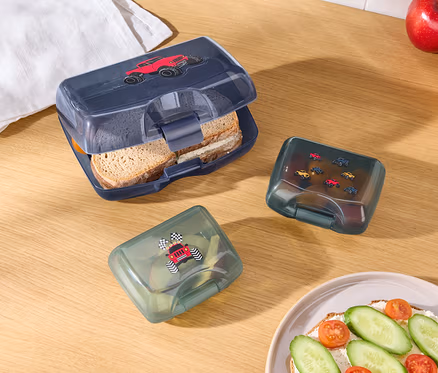 Sur une table en bois, il y a une boîte à goûter bleue ouverte avec une voiture rouge dessus, deux boîtes à goûter vertes avec des images de voitures, une pomme, une orange, un torchon blanc et une assiette avec une tartine.