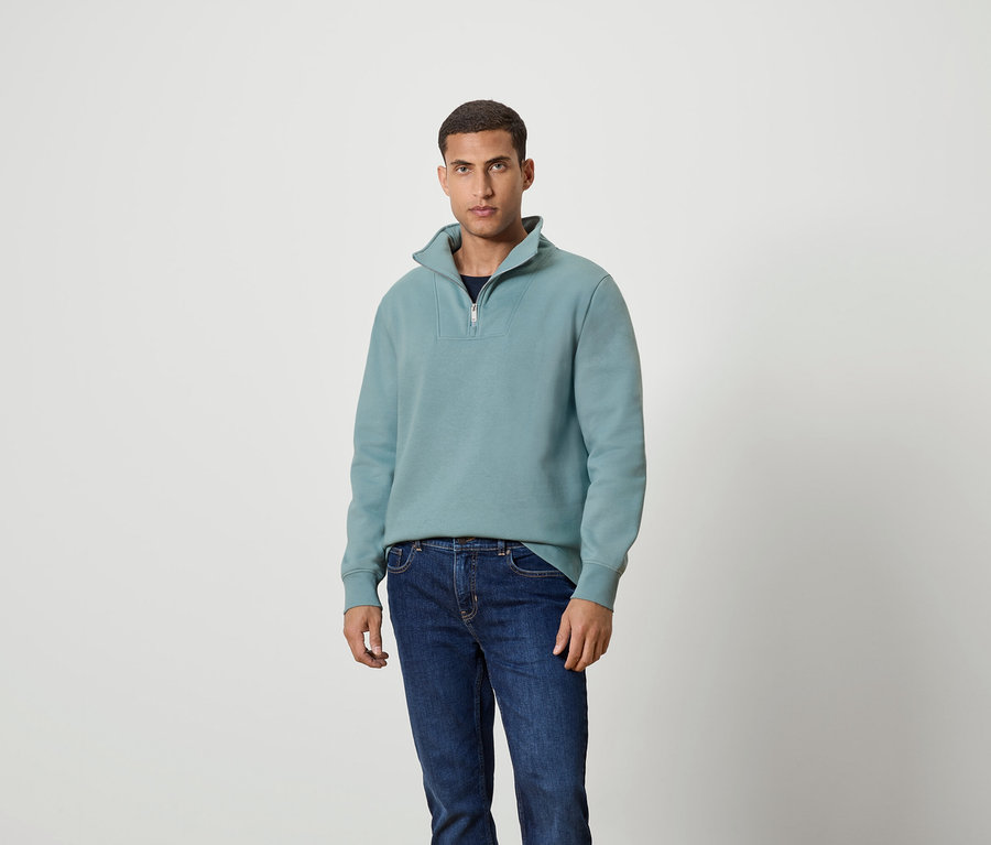 Un homme debout portant un sweat-shirt turquoise à fermeture éclair et un jean.