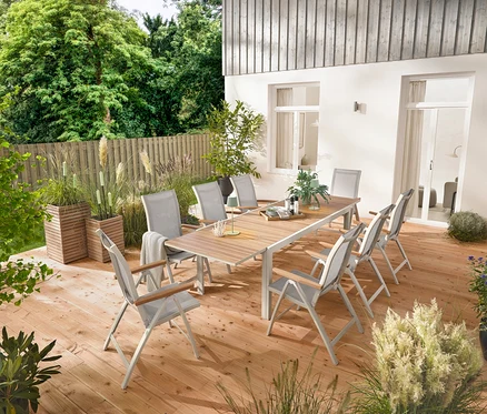 Une table de jardin extensible en aluminium « Liska » d'environ 200 à 300 cm de long est installée sur une terrasse en bois, entourée de six fauteuils à dossier haut « Liska » avec dossier réglable. Deux colonnes à plantes « Tinus » et cinq torches de jardin à LED sont également visibles.