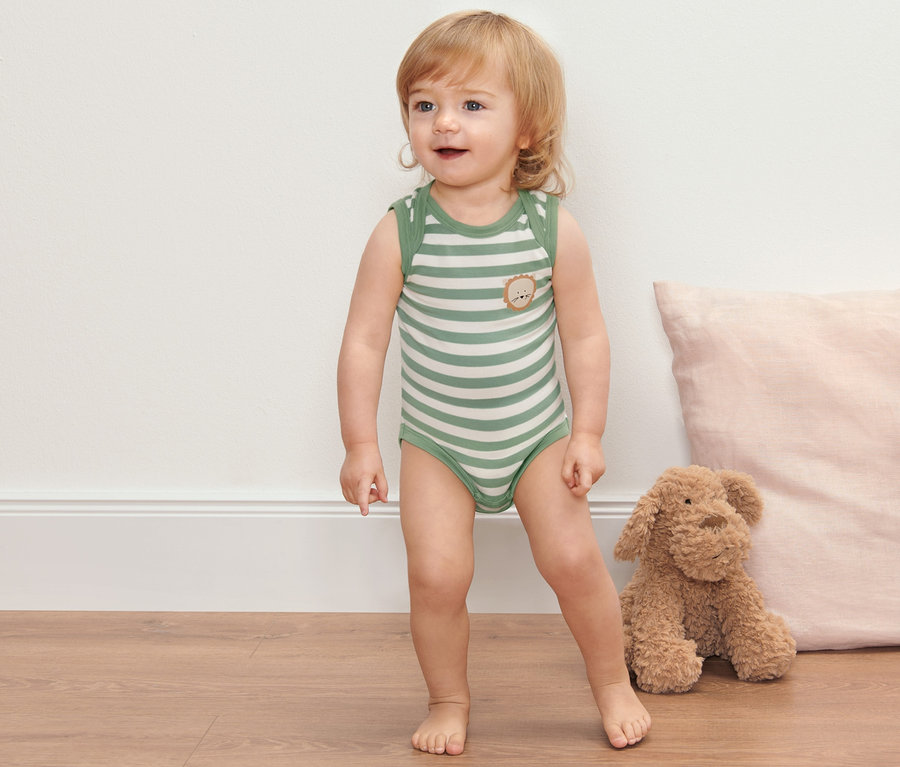 Un bébé se tient debout sur le sol et porte 2 bodys pour bébé avec fonction évolutive.