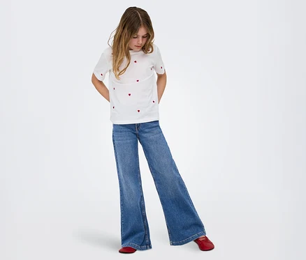 Une jeune fille aux longs cheveux bruns, vêtue d'un t-shirt blanc à motifs de cœurs rouges, d'un jean bleu large et de chaussures plates rouges, regarde vers le bas.