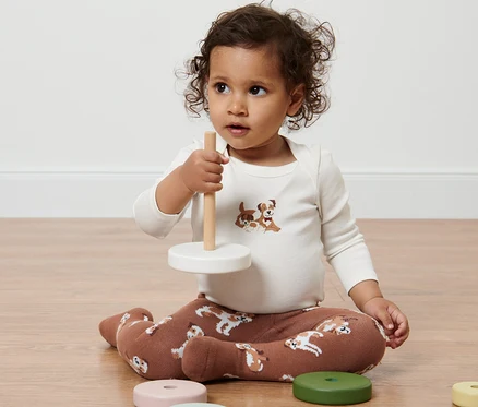 Un enfant est assis par terre et joue avec un jouet. Il porte 2 collants pour bébés avec un motif de chien.