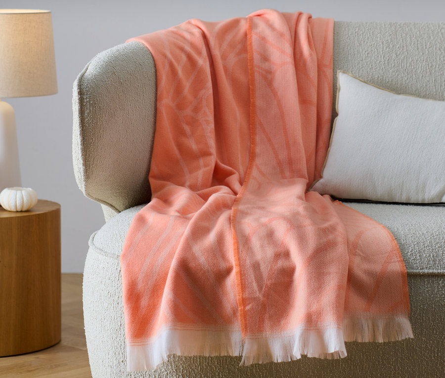Une couverture de salon réversible orange à franges est posée sur un canapé beige à côté d'un coussin blanc.