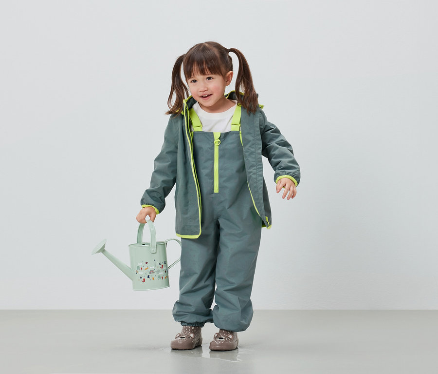 Fillette en pantalon et veste de pluie gris tenant un arrosoir vert clair.