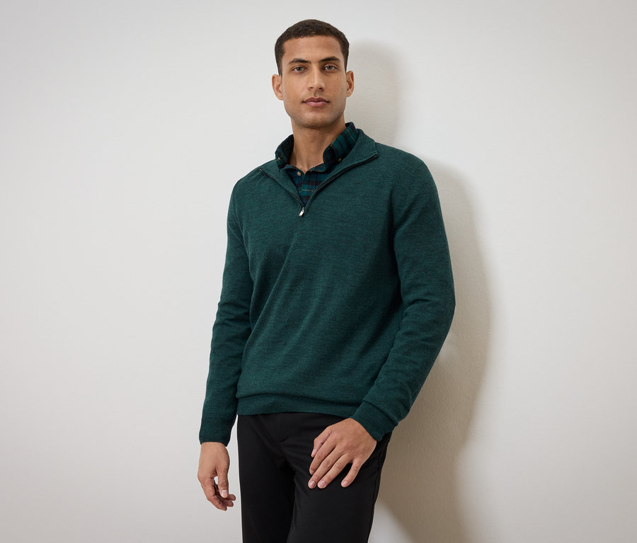 Un homme pose en pull vert et pantalon noir.
