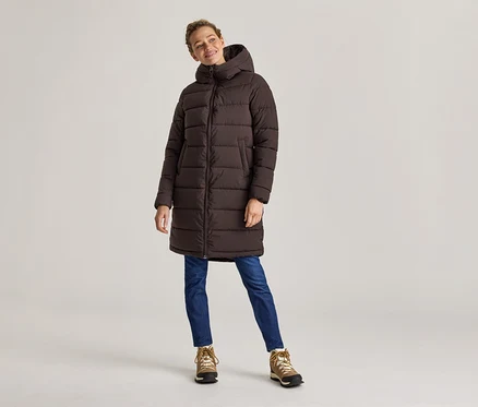 Une femme en manteau bouffant long marron, jean et chaussures de randonnée se tient devant un fond clair.