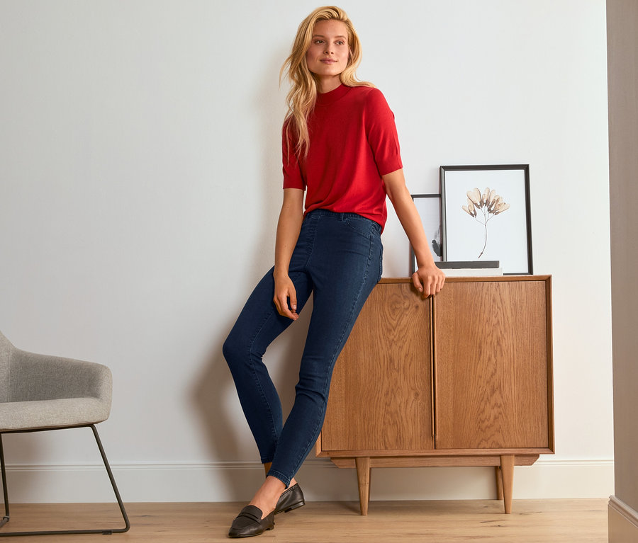 Femme blonde en pull rouge et tregging bleu appuyée contre un meuble en bois.