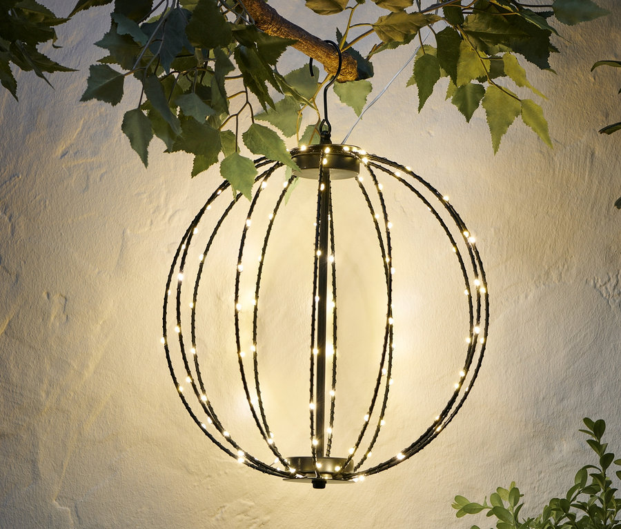 Une sphère LED en métal est suspendue à une branche devant un mur blanc.