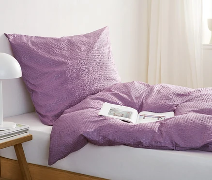 Parure de lit violette en seersucker sur un lit avec un livre ouvert. Une lampe et des livres sont posés sur la table de chevet.