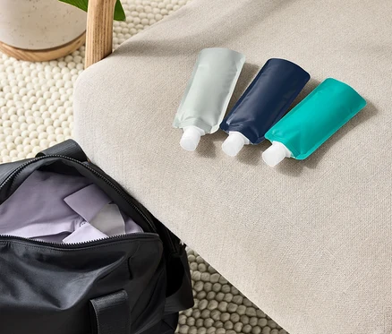 Trois bouteilles de voyage réutilisables sont posées sur un fauteuil beige à côté d'un sac de sport noir et d'une plante en pot.