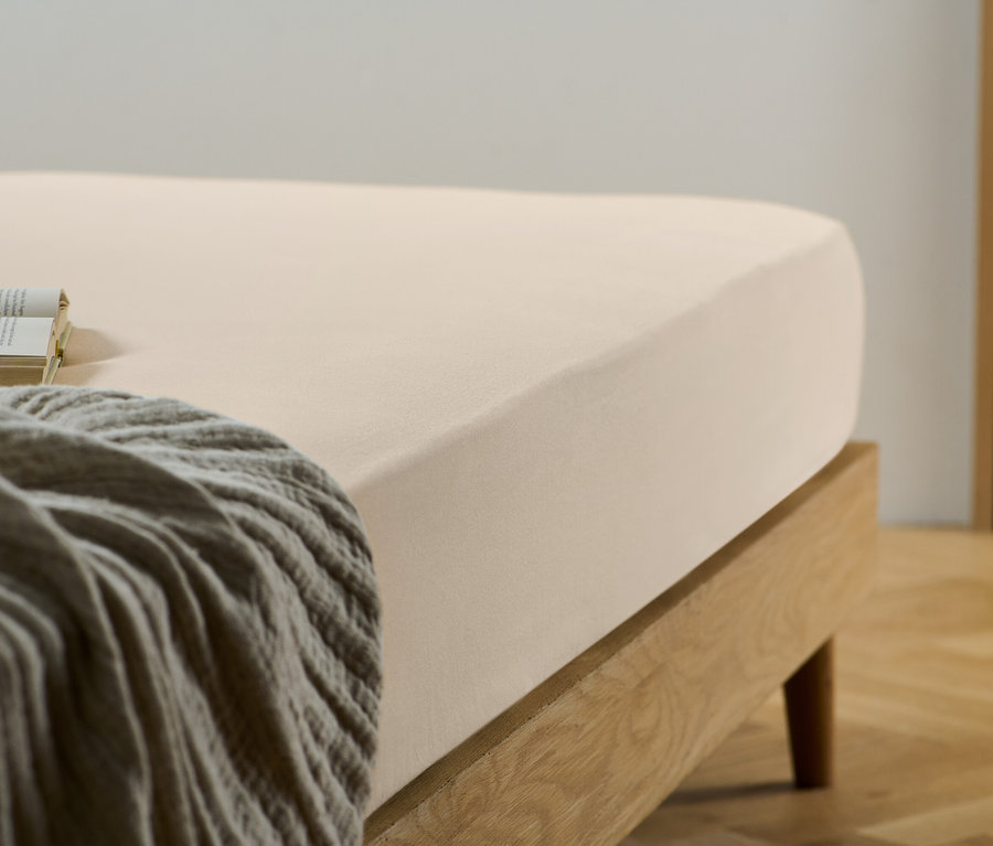 Un drap-housse beige sur un lit avec un cadre en bois. Un livre et une couverture grise sont posés sur le lit.