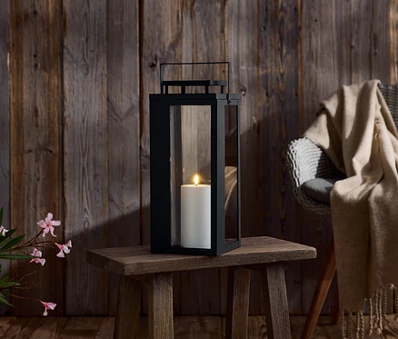 Une lanterne noire avec une bougie blanche sur une petite table en bois. En arrière-plan, un mur en bois et une chaise avec une couverture.