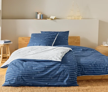 Lit avec des draps rayés bleu et blanc et un motif de nœuds. Sur la table de chevet, il y a des livres et une lampe en forme de bateau.
