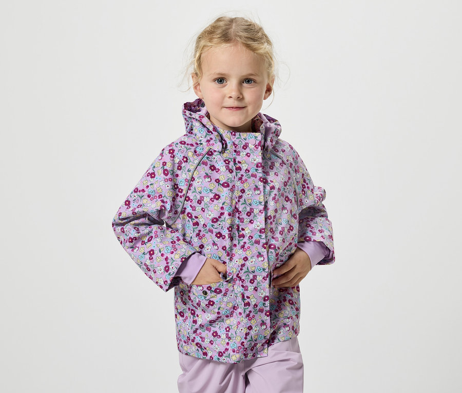 Une fillette blonde portant une veste à fleurs lilas se tient devant un fond blanc.