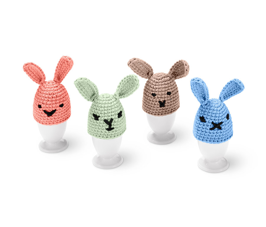 Quatre couvre-œufs crochetés en forme de lapin de couleurs saumon, vert, beige et bleu sont posés sur des coquetiers blancs. Kit DIY pour couvre-œufs en crochet.