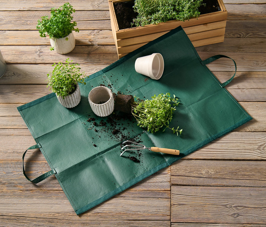 Un sac à déchets de jardin repose sur un plancher en bois à côté de plantes en pot et d'un arrosoir.