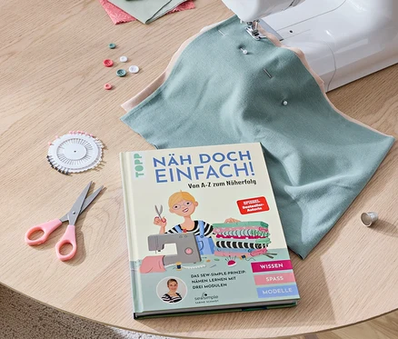Sur une table se trouvent une machine à coudre, le livre « Näh doch einfach », des tissus, des ciseaux, des boutons et des épingles.