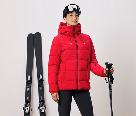 Femme en veste de ski rouge tenant des bâtons, skis à côté et lunettes de ski sur la tête.