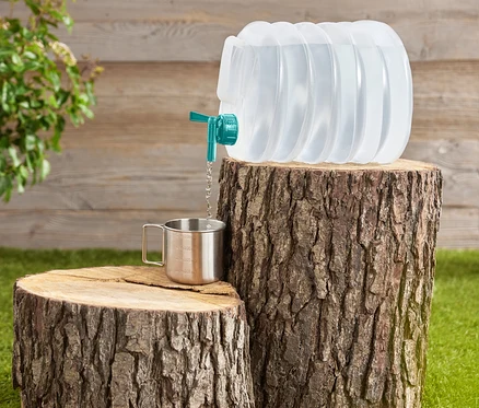 L'eau est versée d'un récipient d'eau pliable et transparent avec un robinet turquoise dans une tasse en acier inoxydable placée sur des souches d'arbres.