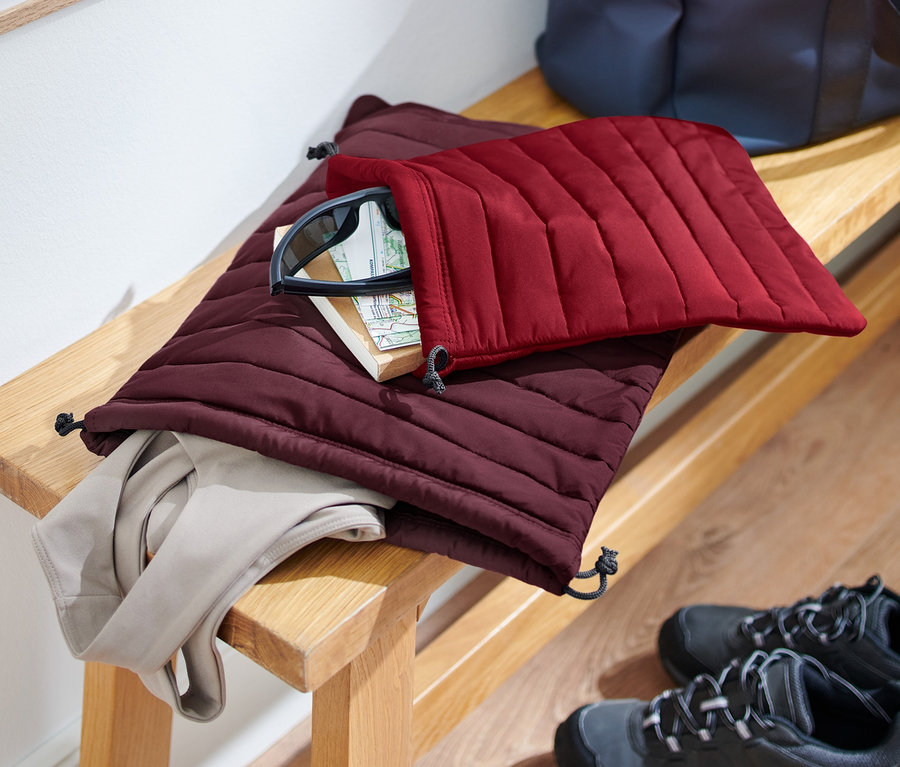 Sur un banc, 2 organisateurs matelassés, un bordeaux et un rouge foncé, avec du contenu. Des chaussures sont posées sur le sol.