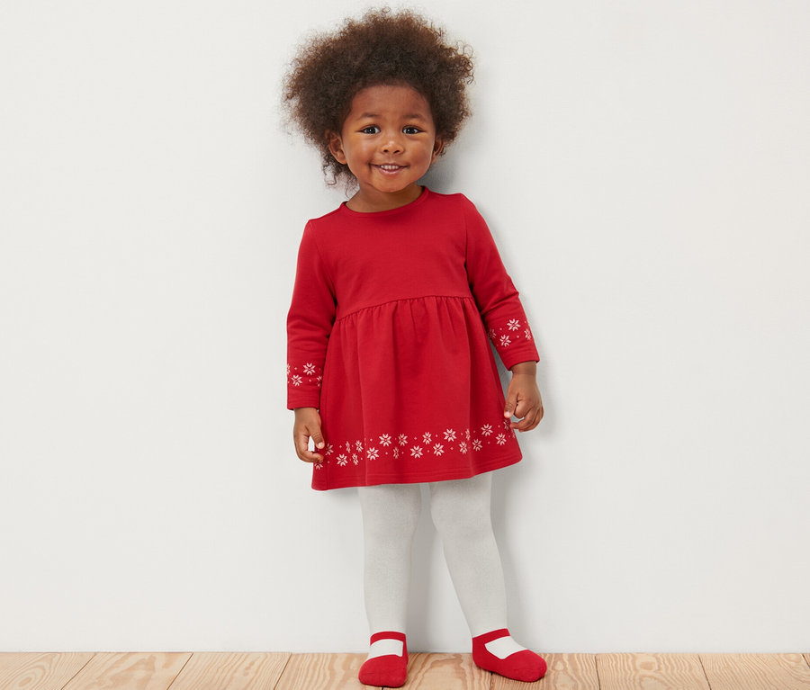 Une petite fille souriante dans une robe rouge avec des flocons de neige se tient sur un plancher en bois et porte des collants blancs et des chaussures rouges. Elle porte aussi 2 collants thermiques pour bébé, ballerine.