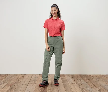 Une femme se tient devant un mur, portant une chemise rouge à manches courtes et un pantalon vert.